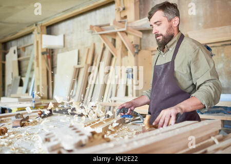 Portrait of mature carpenter barbu faire des meubles en atelier moderne, soufflant au large de copeaux de Workstation, copy space Banque D'Images