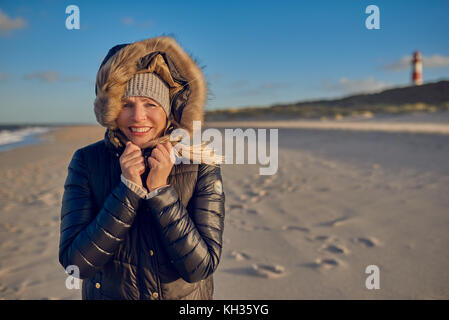 Portrait of a Beautiful woman smiling, tout en portant un manteau d'hiver à capuchon étanche noir sur une plage vide avec un vieux phare à l'horizon Banque D'Images