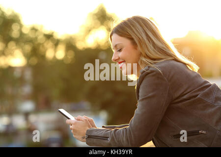 Vue latérale d'une femme heureuse en utilisant un téléphone intelligent dans un foyer balcon au coucher du soleil Banque D'Images