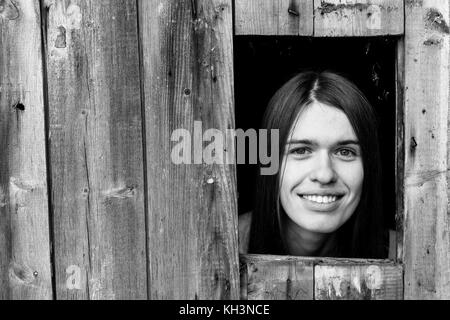 Une jeune femme enfermée dans une cabane en bois, peeking through une petite fenêtre. noir et blanc photo. Banque D'Images