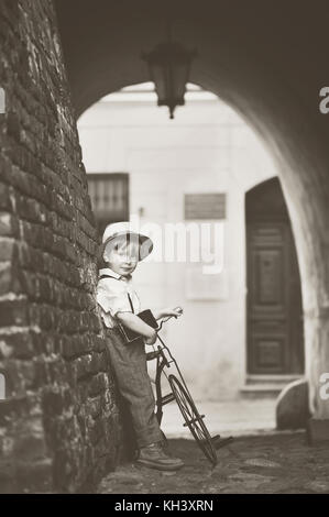 Un petit garçon souriant habillé en style retro debout dans l'allée de la vieille ville et la tenue d'une bicyclette. Noir blanc. Young boy leaning against wall Banque D'Images