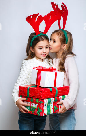 Deux happy little smiling girls avec cadeau de noël de ticket..concept de Noël. Smiling funny sœurs en cornes de cerf en studio de dire à un secret. Banque D'Images