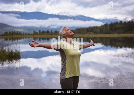 Happy senior woman with arms outstretched standing près du lac Banque D'Images