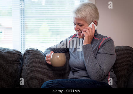 Senior Woman talking on mobilephone tandis que boire du café à la maison Banque D'Images