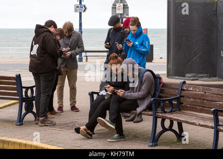 Groupe de jeunes / étudiants absorbés dans leurs téléphones portables sur le front de mer à Aberystwyth Banque D'Images