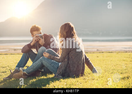 Jeunes amis assis sur l'herbe, il prend une photo de sa petite amie à l'aide d'un appareil photo numérique, sur fond de paysage naturel Banque D'Images