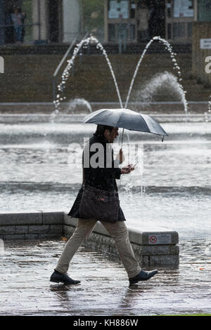 Homme marchant dans les fortes pluies, holding umbrella & utilisation du téléphone, l'adoption de la piscine miroir et fontaines - Bradford City Park, West Yorkshire, Angleterre, Royaume-Uni. Banque D'Images