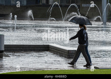 Couple en train de marcher dans de fortes pluies, s'abritant sous un parapluie, l'adoption de la piscine miroir et fontaines - centre de Bradford City Park, West Yorkshire, Angleterre, Royaume-Uni. Banque D'Images