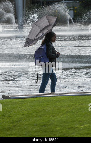 Femme marche dans la pluie et le vent, avec brolly intérieur à l'extérieur, en passant par la piscine miroir et fontaines - Bradford City Park, West Yorkshire, Angleterre, Royaume-Uni. Banque D'Images