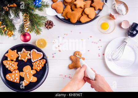 Femme mains tenant un sac avec de la crème culinaire meringue inprocess de décorer gingerbread man cookies. table en bois blanc avec ingrediens et décoration de détails. fond de noël concept. Banque D'Images