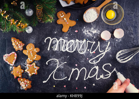 Vue de dessus de la Crème mains femme meringue lettrage joyeux noël sur la pierre noire avec fond ingrediens et décoration. détails Maison de Vacances Nouvel an concept. la cuisine et le processus de décoration. Banque D'Images