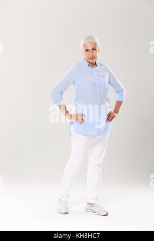 Photo de pleine longueur senior sérieux femme aux cheveux gris, debout avec les mains sur sa taille, looking at camera, isolé sur fond blanc Banque D'Images