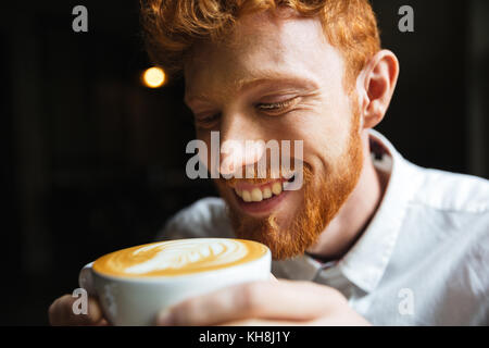 Close-up portrait of smiling curly readhead barbu dans la tasse de café de dégustation Banque D'Images