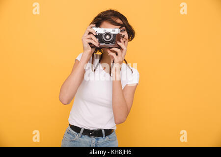 Portrait d'une jeune fille brune de prendre une photo avec un appareil photo rétro en position debout sur fond jaune isolé Banque D'Images