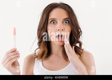 Choqué brunette woman holding test de grossesse positif et regardant la caméra sur fond blanc Banque D'Images