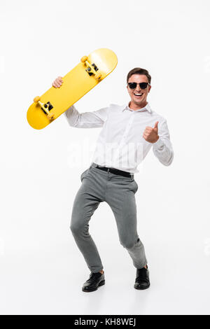 Portrait d'un homme heureux succès en chemise blanche et lunettes posant avec une planche à roulettes et showing Thumbs up tout en geste et permanent Banque D'Images