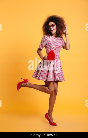 Longueur totale de la photo jolie femme élégante rétro africaine en robe et talons hauts qui posent avec big red lips, isolé sur fond jaune Banque D'Images