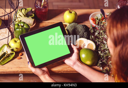 Femme montrant une tablette avec un écran vert à la table de la cuisine avec des légumes verts et des fruits, le chroma copyspace Banque D'Images