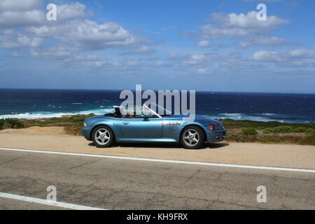 Une jeune femme heureuse assise dans une voiture de sport cabrio cabrio convertible, vue panoramique sur la plage en arrière-plan. Banque D'Images