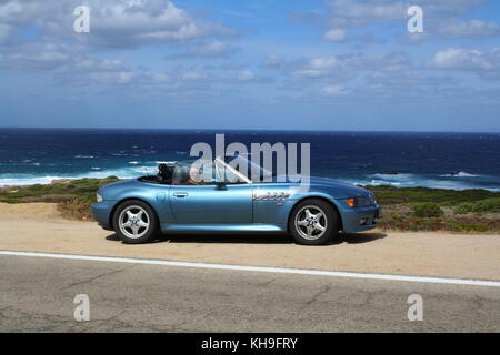 Une jeune femme heureuse assise dans une voiture de sport cabrio cabrio convertible, vue panoramique sur la plage en arrière-plan. Banque D'Images