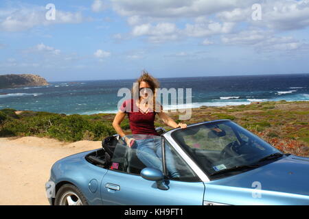 Une jeune femme heureuse assise dans une voiture de sport cabrio cabrio convertible, vue panoramique sur la plage en arrière-plan. Banque D'Images