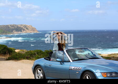 Une jeune femme heureuse assise dans une voiture de sport cabrio cabrio convertible, vue panoramique sur la plage en arrière-plan. Banque D'Images