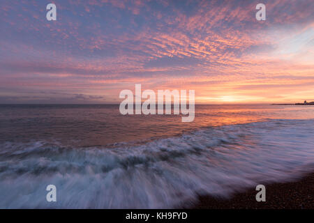 Novembre un coucher du soleil à Hastings, East Sussex, UK avec un ciel rose et orange et des vagues sur la plage de galets. Le soleil se reflète dans l'eau. Banque D'Images