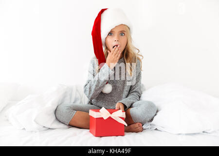 Petite fille a surpris à Santa's hat holding present alors qu'il était assis les jambes croisées au lit Banque D'Images