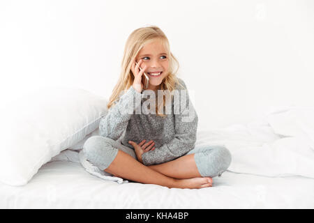 Happy little girl en pyjama gris en conversation sur smartphone, à côté tandis que sitting on bed Banque D'Images