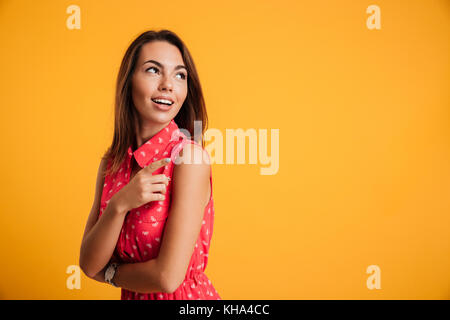 Portrait de jeune femme brune assez étonné en robe rouge pointant avec le doigt, à côté, isolé sur fond jaune