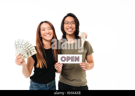 Portrait of a smiling young couple gai montrant tas de billets d'argent et d'un open sign isolated over white background Banque D'Images