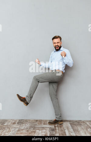 Longueur totale image of screaming barbu en vêtements d'affaires se réjouissent et regardant la caméra sur fond gris Banque D'Images