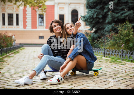 Deux heureux jeunes adolescentes doigt tout en étant assis sur une planche à roulettes sur une rue de la ville en plein air Banque D'Images