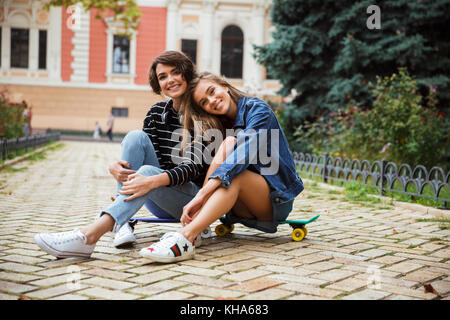 Deux jeunes adolescentes souriant assis sur une planche à roulettes et à la recherche à l'appareil photo sur une rue de la ville en plein air Banque D'Images