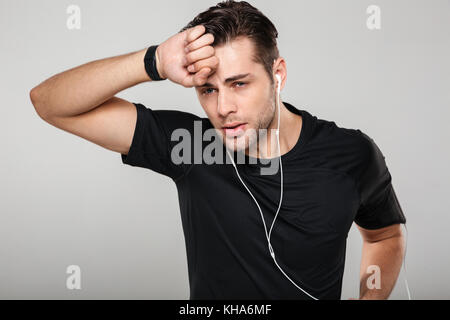 Portrait d'un jeune sportif en sueur fatigué en essuyant son front écouteurs sur fond gris isolé Banque D'Images