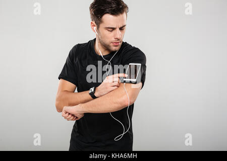 Portrait d'un beau jeune homme sportif des écouteurs d'écouter de la musique en touchant son téléphone en isolé sur fond gris brassard Banque D'Images