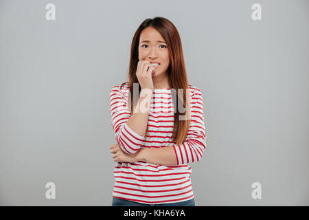 Portrait d'une fille asiatique nerveux de mordre ses ongles tout en regardant plus isolé de l'appareil photo arrière-plan gris Banque D'Images