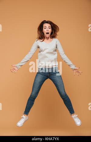 Photo de pleine longueur choqué jeune femme en blouse grise et un jean saute au-dessus de fond beige Banque D'Images