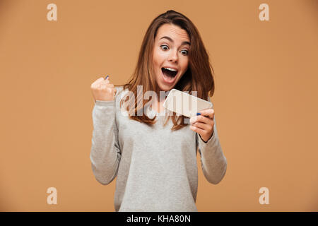 Happy attractive brunette girl joying gagner en jeu vidéo sur smartphone, isolé sur fond beige Banque D'Images