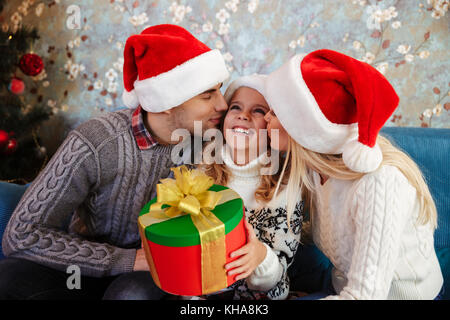 Jeune père et mère à Santa's hat embrassant ces petite fille, assise sur un canapé bleu au matin de Noël Banque D'Images