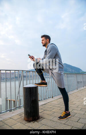 Un beau jeune homme mettant sa jambe vers le haut et se penchant sur elle tout en utilisant son téléphone du riverside Banque D'Images