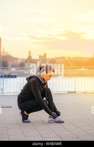 Une belle jeune femme attachant sa chaussure dans le coucher du soleil au bord de la rivière tandis que crouching Banque D'Images