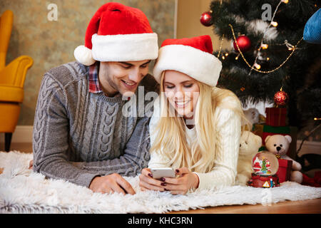 Happy young couple in Santa's hat looking at mobile phone en position couchée à l'étage Banque D'Images