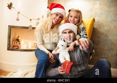 Young smiling père dans Santa's hat pointant avec le doigt sur vous, holding gift box alors qu'il était assis avec sa famille à la cheminée Banque D'Images