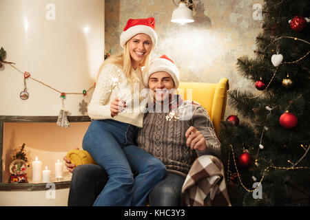 Jeune couple charmant à Santa's hat holding sparklers, looking at camera while sitting in chair Banque D'Images