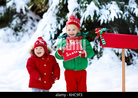 Heureux les enfants en renne tricoté bonnet et écharpe holding lettre au père Noël avec des cadeaux de Noël liste de souhaits au red boîte de neige sous arbre de Noël dans Banque D'Images