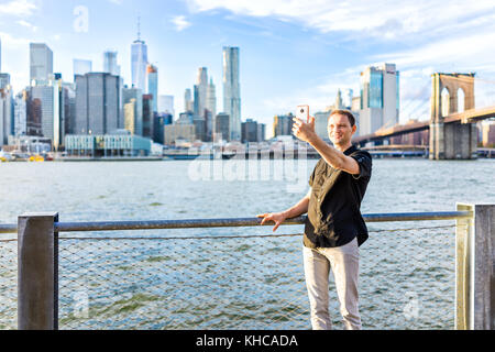 Jeune hipster homme debout en tenant photo selfies par une clôture à Brooklyn Bridge Park surplombant le Nyc New York City Manhattan cityscape skyline avec w Banque D'Images