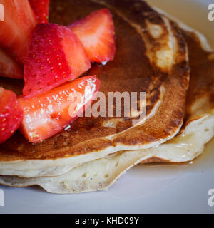 Gros plan d'une pile de crêpes maison sur une plaque blanche avec des fraises coupées et sirop d'érable sur le dessus. Format carré. Banque D'Images