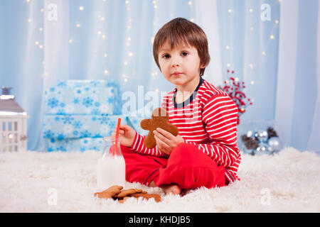 Adorable petit garçon heureux, manger des cookies et boire du lait, en attente de Santa dans la veille de Noël nuit pyjama Banque D'Images