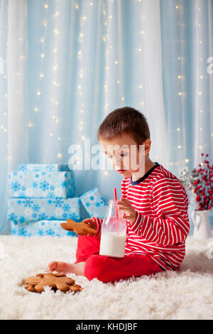 Adorable petit garçon heureux, manger des cookies et boire du lait, en attente de Santa dans la veille de Noël nuit pyjama Banque D'Images